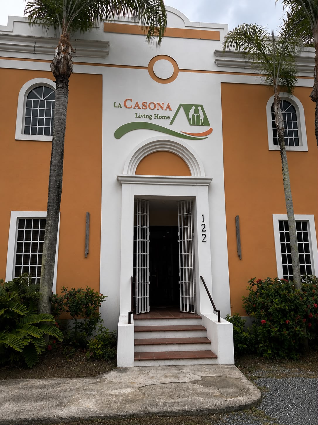 Entrada de La Casona Living Home con su logo sobre la puerta de arco, escaleras y palmeras